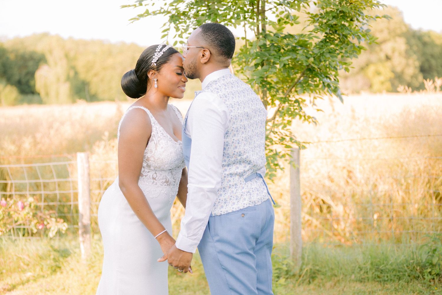 A couple shares a tender moment in a sunlit field with golden grass and trees during a summer wedding celebration.