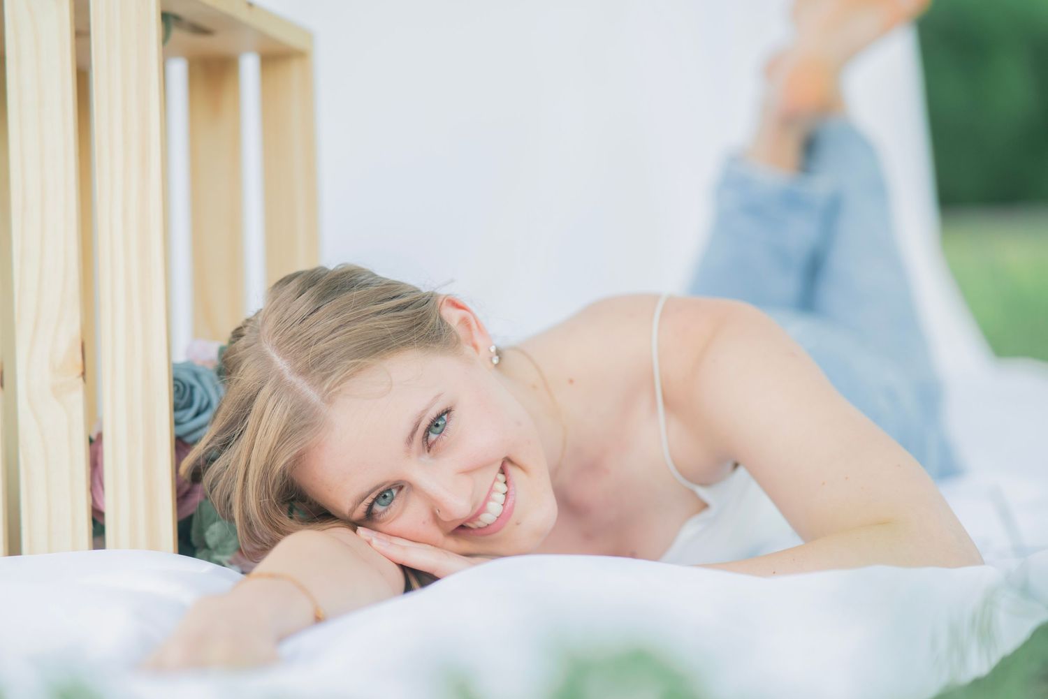 A smiling young blonde woman lies on a white bed outdoors, leaning against a wooden crate in casual attire.