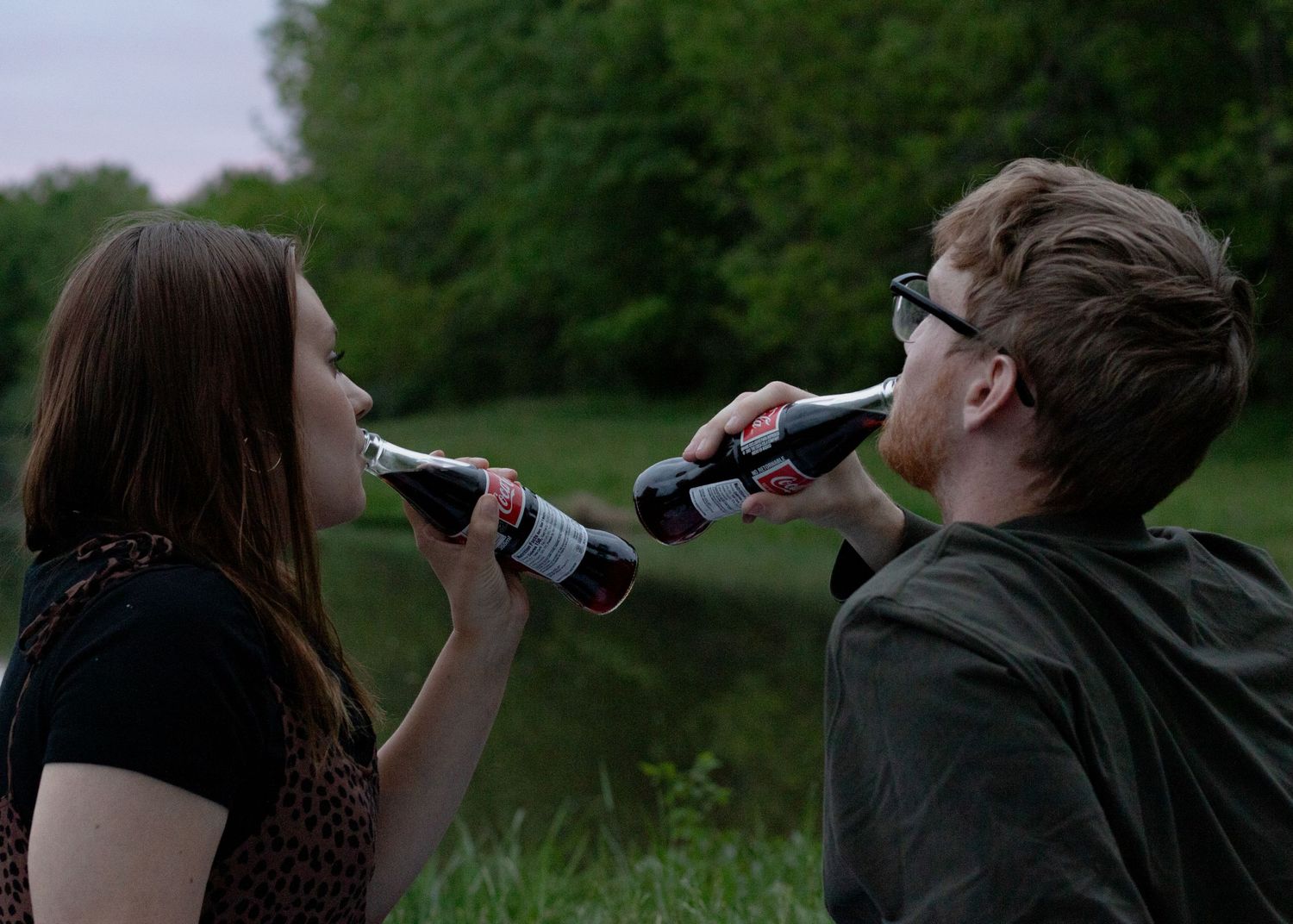 Couple drinking coke from glass bottles in front of a lake at sunset