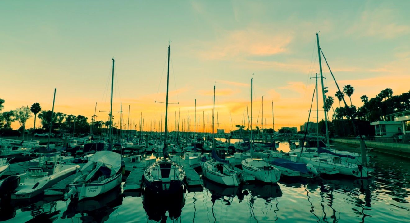 Sailboats with tall masts docked in a calm marina at sunset, with a vivid orange and yellow sky reflecting on the water.