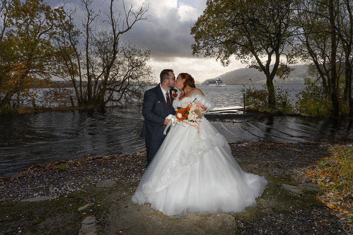 Bride and groom kissing beneath lakeside trees, with her full white skirt and autumn bouquet in view.