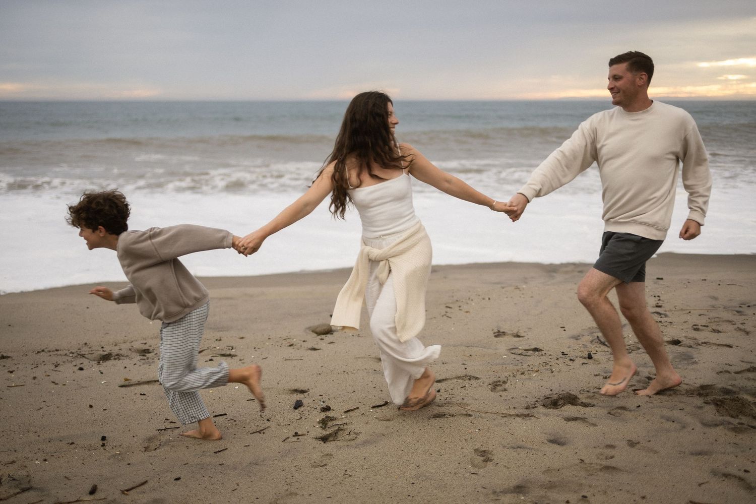 Family of three joyfully running and holding hands on a sandy beach at sunset near ocean waves.