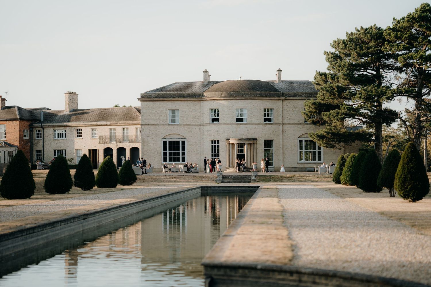 Elegant Georgian country house venue with formal reflecting pool, manicured topiary hedges, and gravel terrace under clear blue sky.