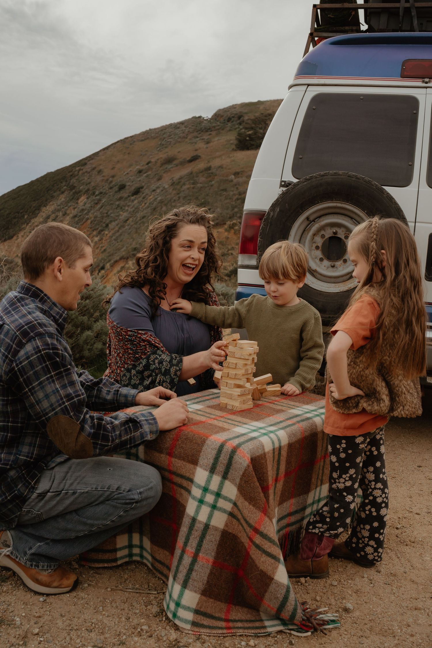 Family sitting in open doors of vintage white and blue van with roof rack on dirt road.