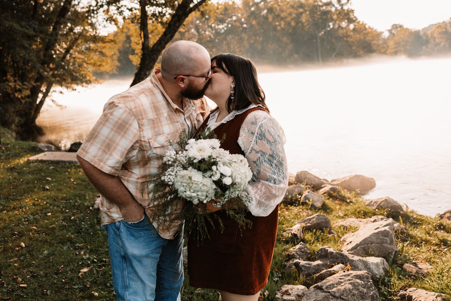 Couple sharing a kiss outdoors by a misty lake, man holding white hydrangea bouquet, wearing casual fall attire.