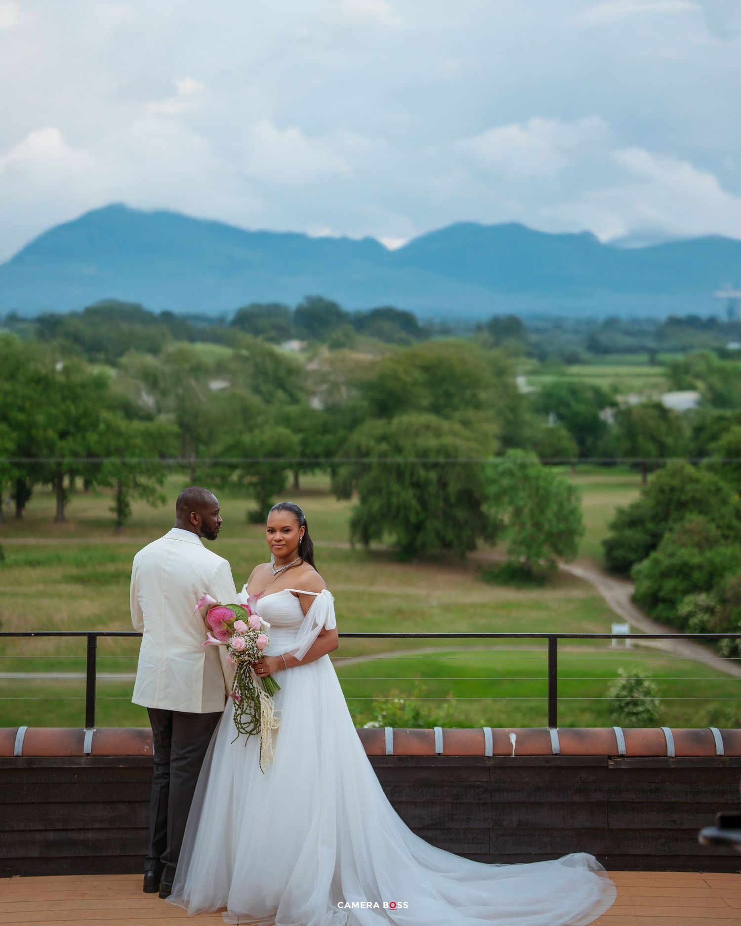Bride and groom share a romantic moment on a terrace with lush green landscape and mountain backdrop.