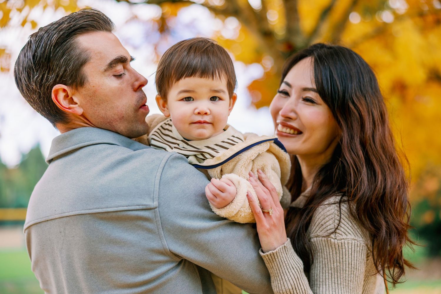 Happy young family with mixed-race baby boy enjoying autumn park, parents kissing and smiling at toddler outdoors.