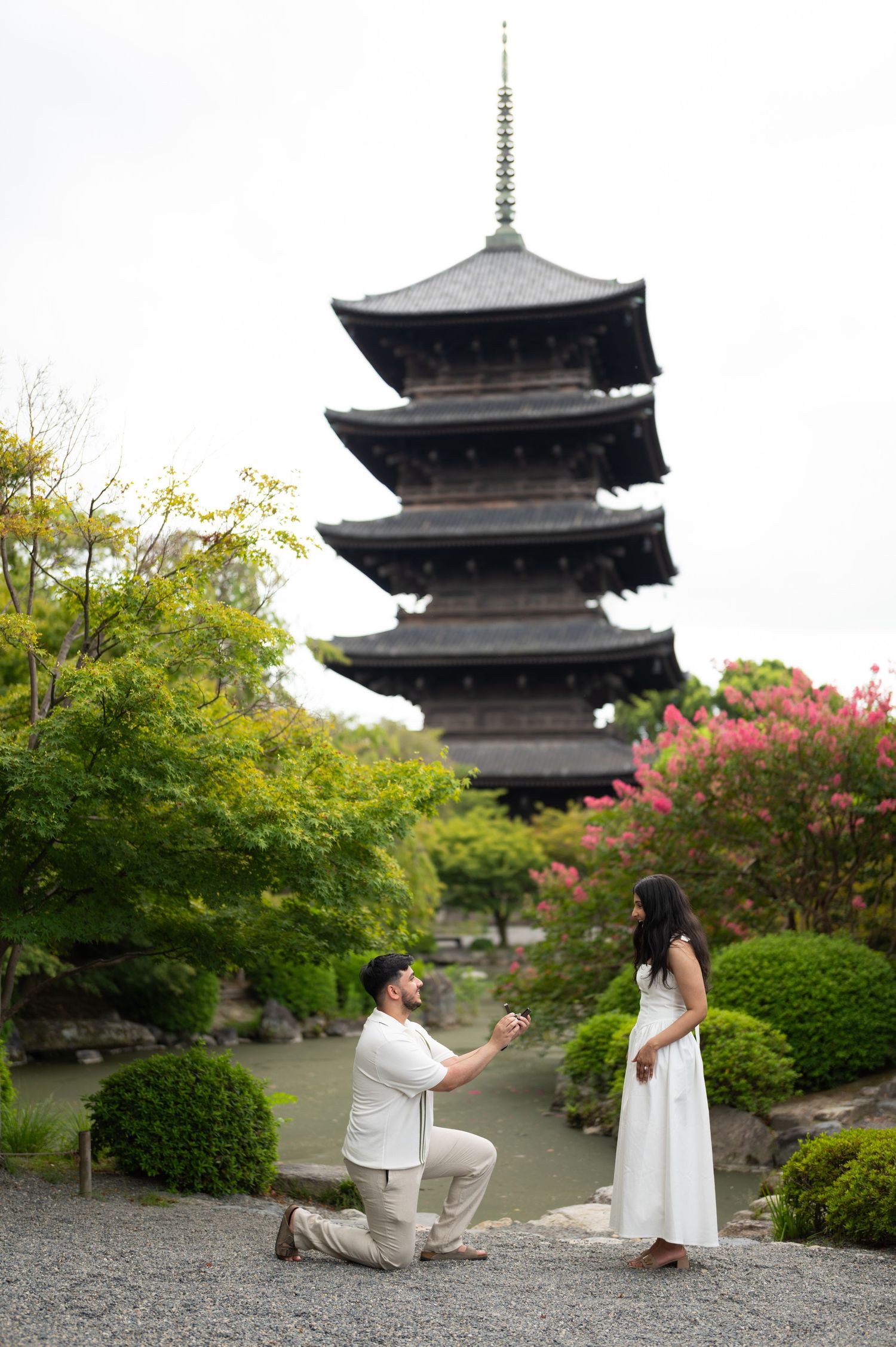 Man kneeling on one knee proposing to a woman in a white dress in a Japanese garden with a five-story pagoda behind them.