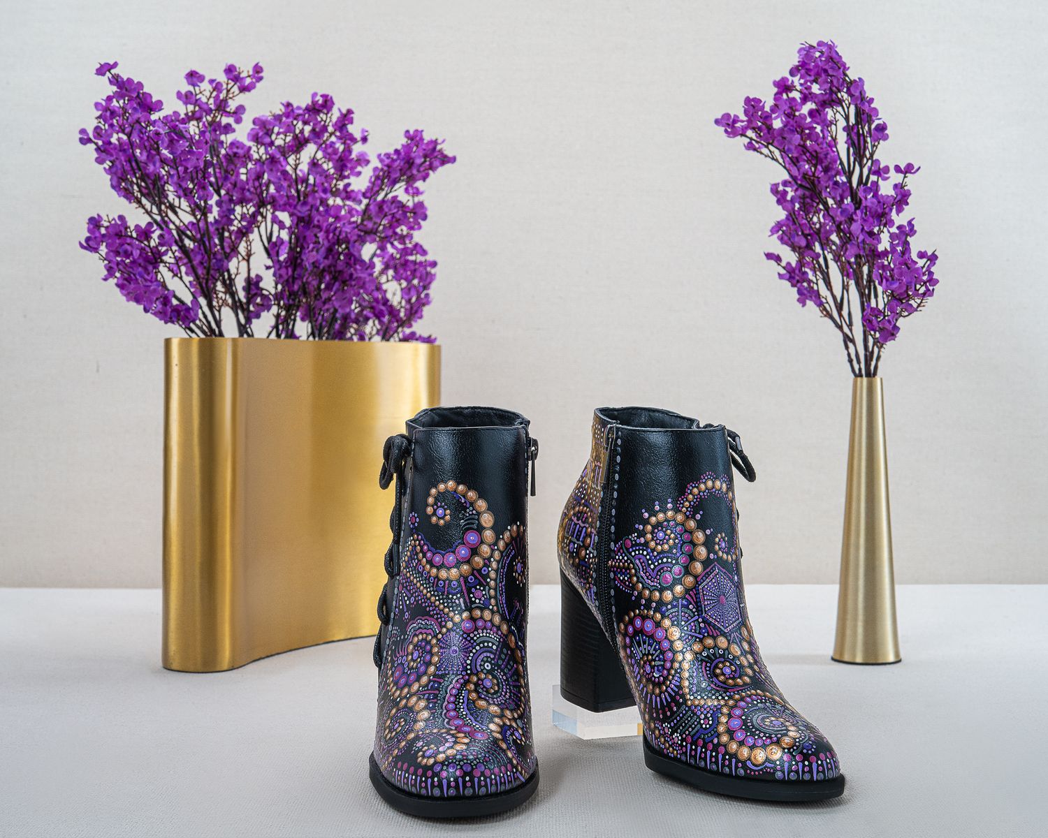 Pair of black block-heel ankle boots with hand-painted purple and gold mandala designs beside gold vases holding purple flowers.
