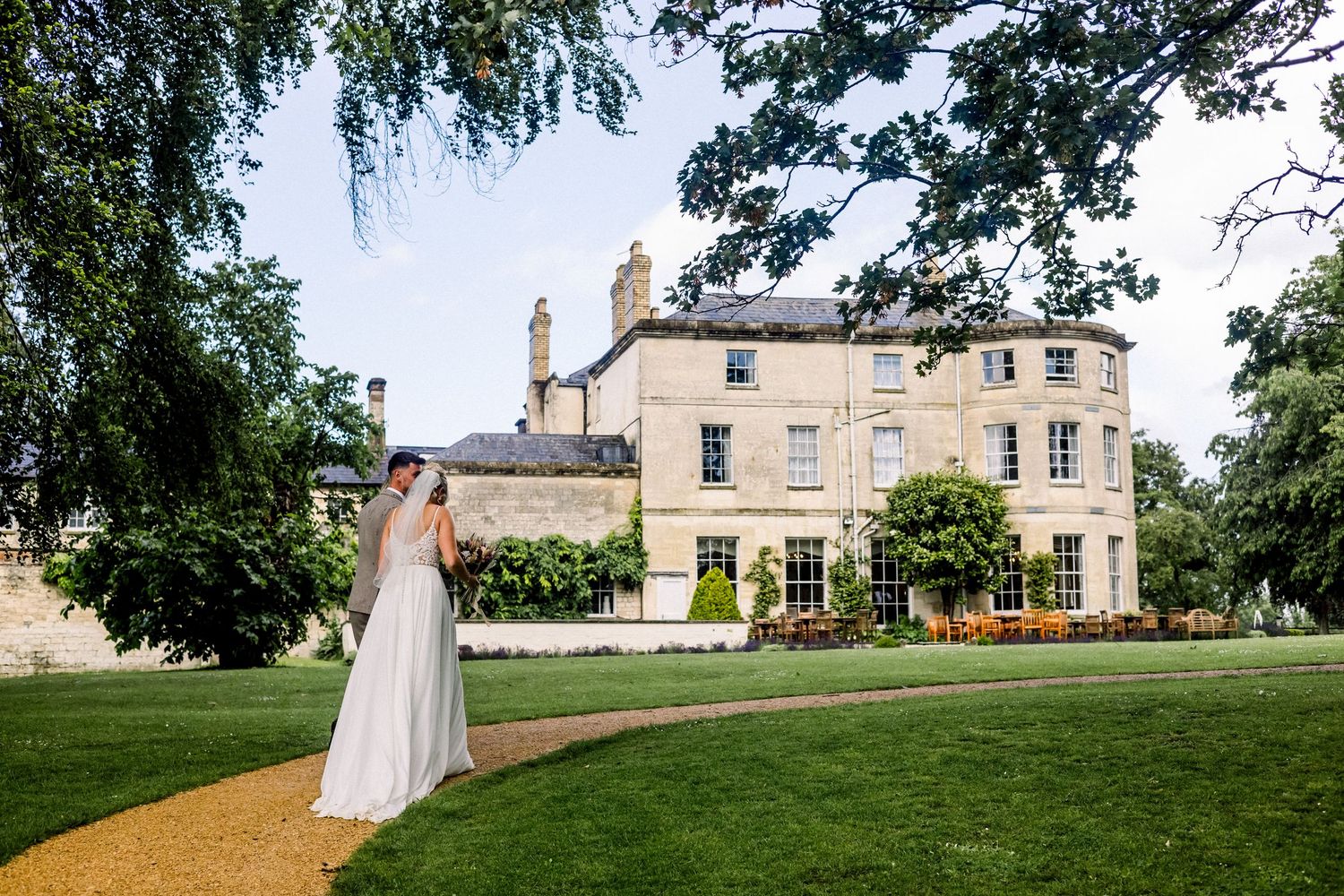Bride in white gown poses on garden path before elegant Georgian country house wedding venue on sunny day.