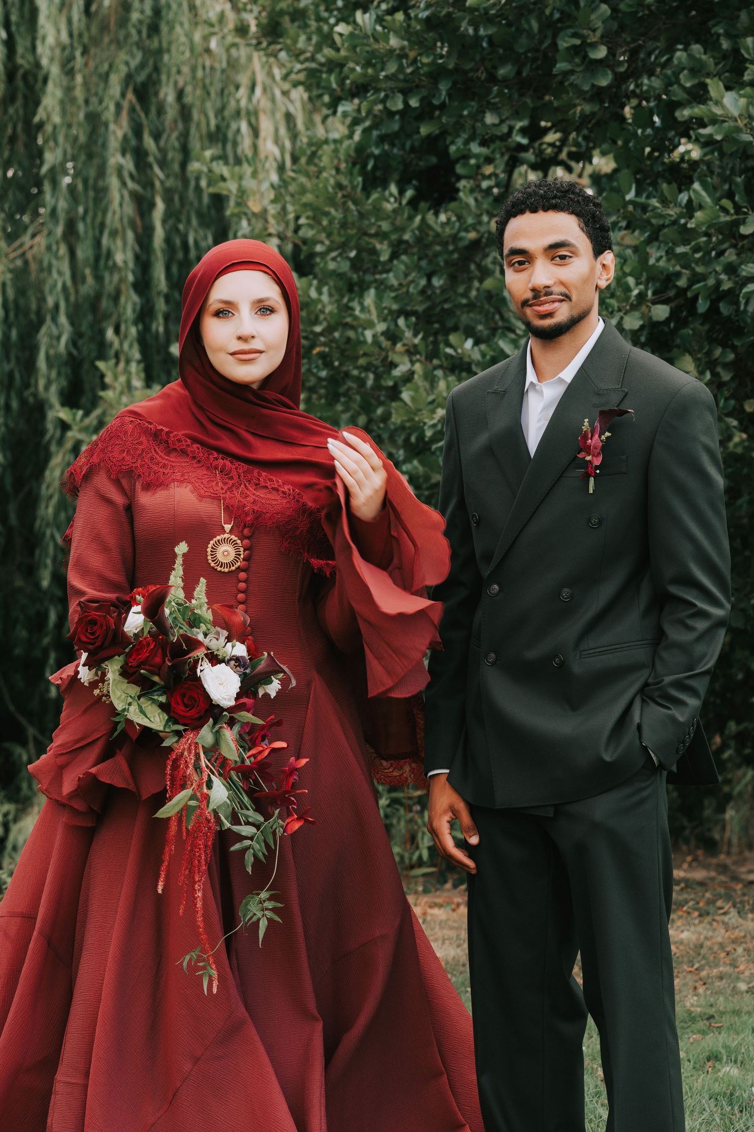 Muslim bride in red hijab and gown holds bouquet beside groom in dark suit with boutonniere outdoors.