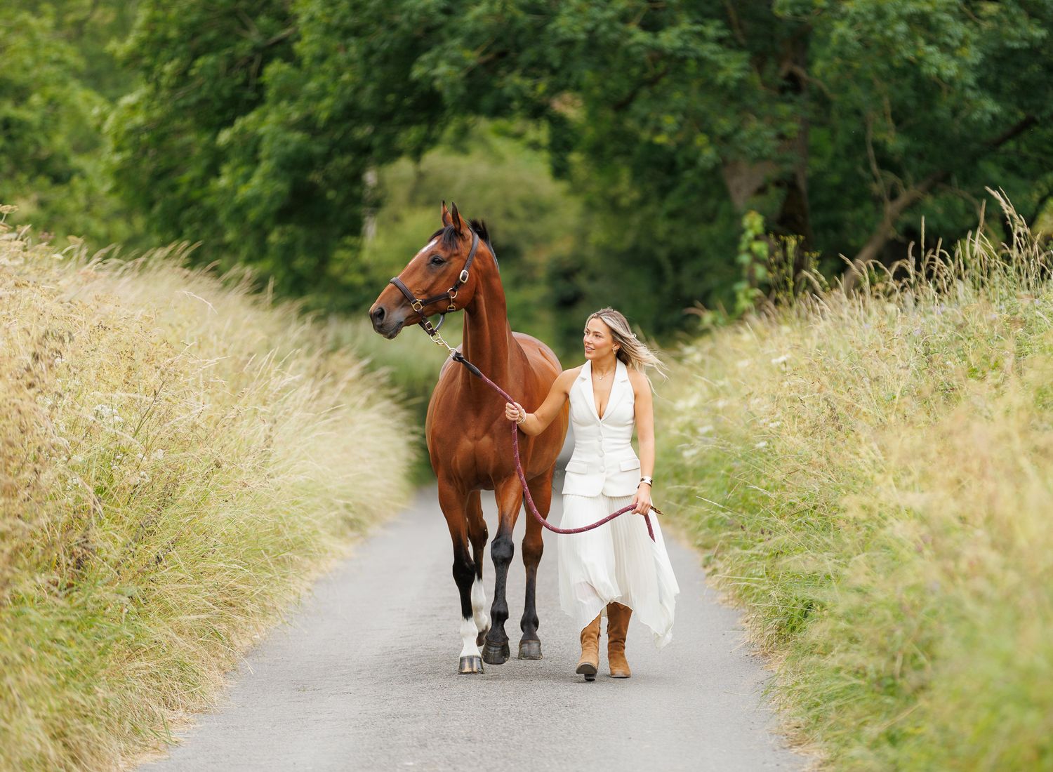 A person leads a palomino horse along a sunlit forest trail surrounded by tall evergreen trees.