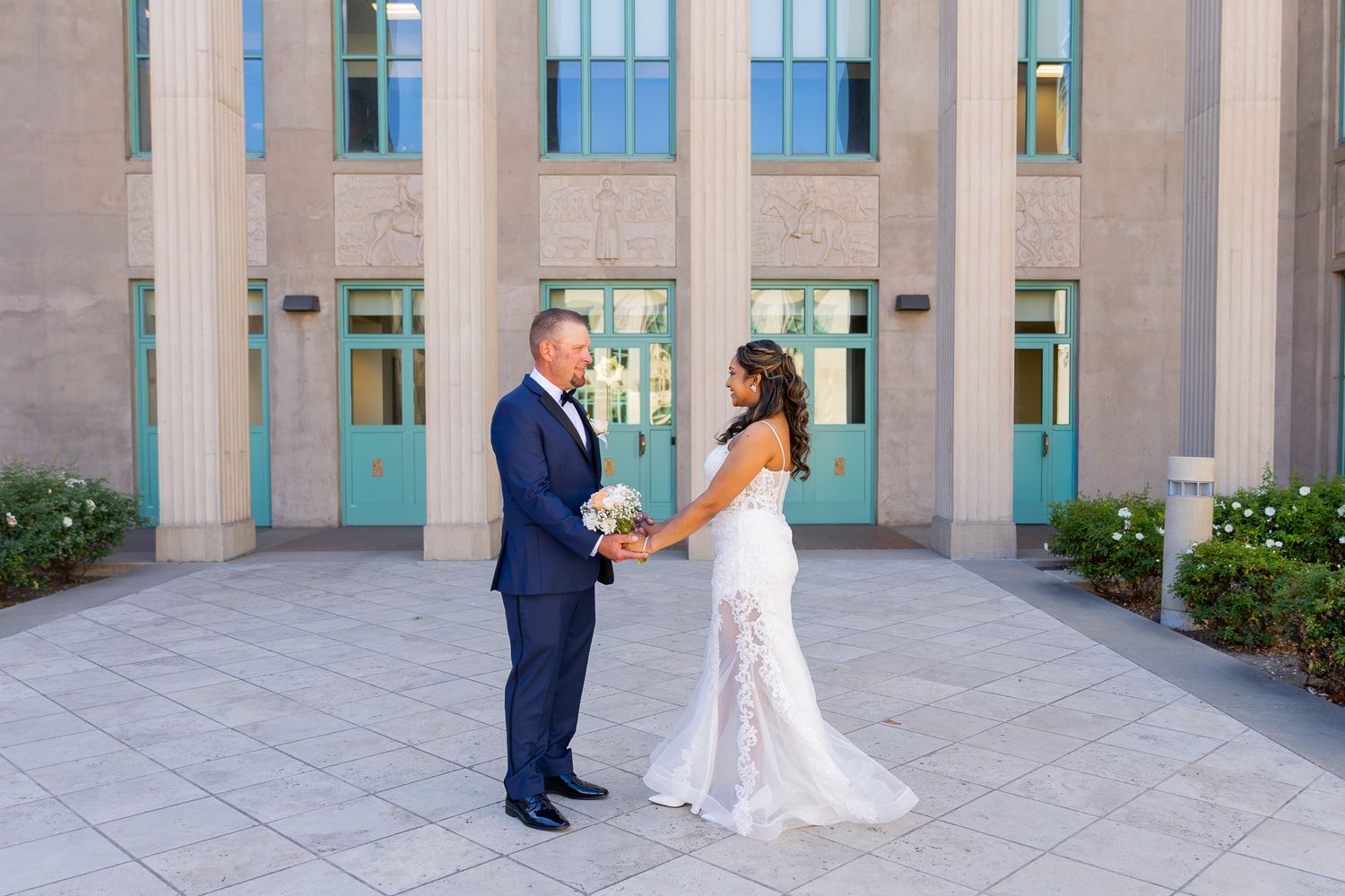 Bride and groom hold hands outside a grand building with teal doors and tall columns on their wedding day.