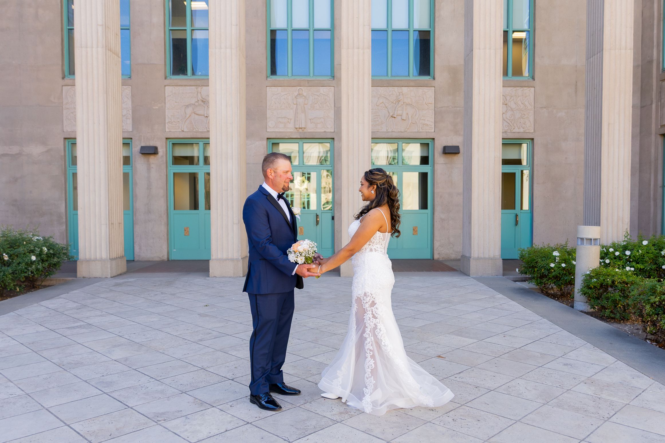Bride and groom hold hands outside a grand building with teal doors and tall columns on their wedding day.