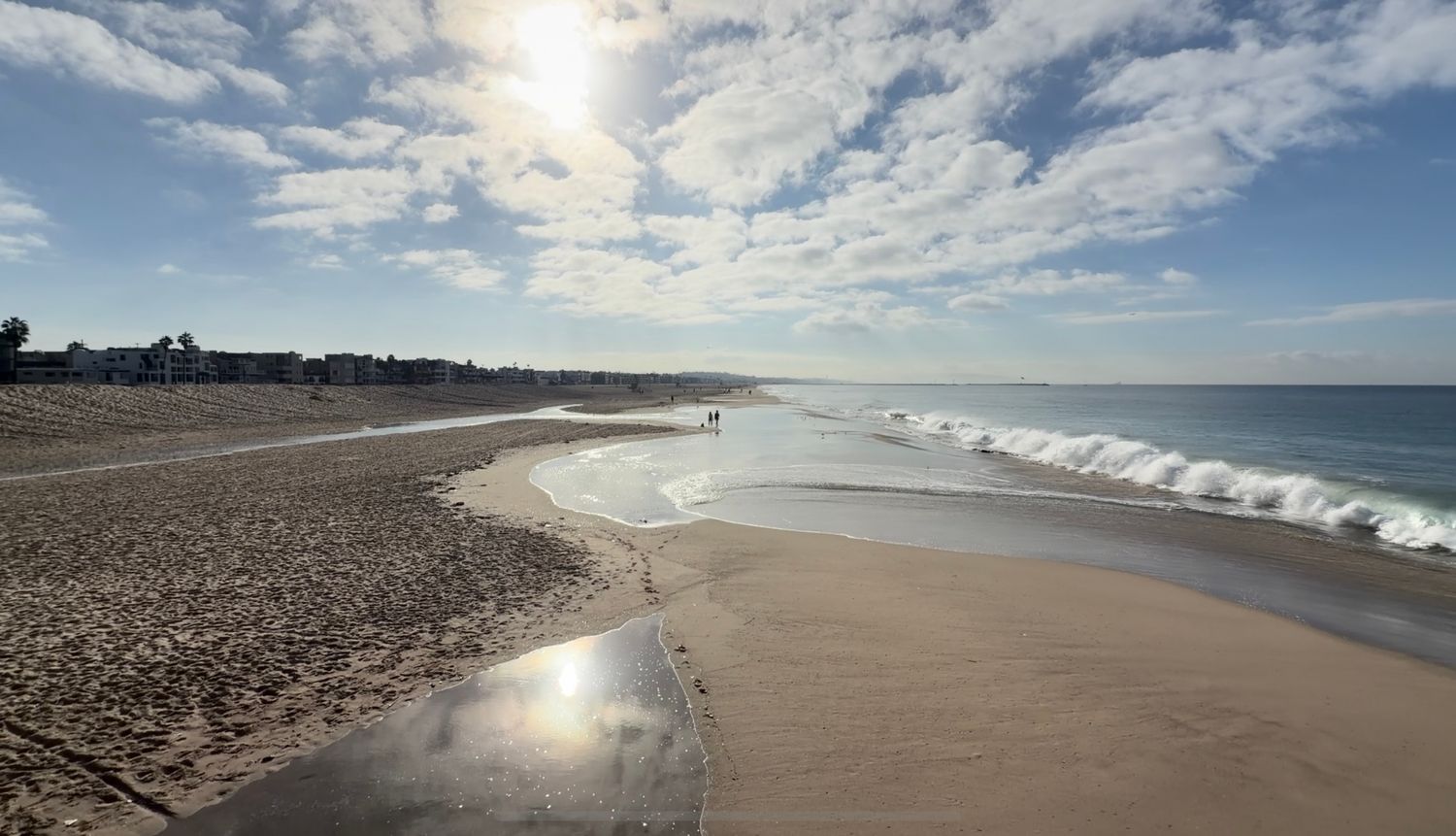 Sunlit sandy beach with gentle waves rolling onto the shore under a bright blue sky with scattered clouds.