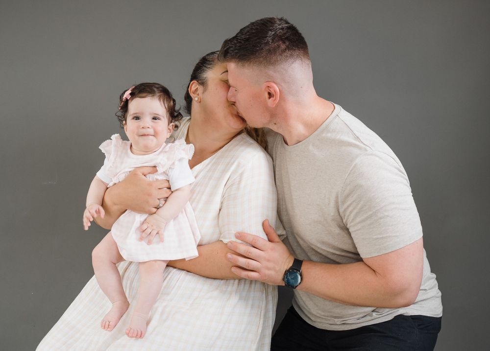 A couple shares a tender kiss while holding their smiling baby girl in a white dress during a studio family portrait.