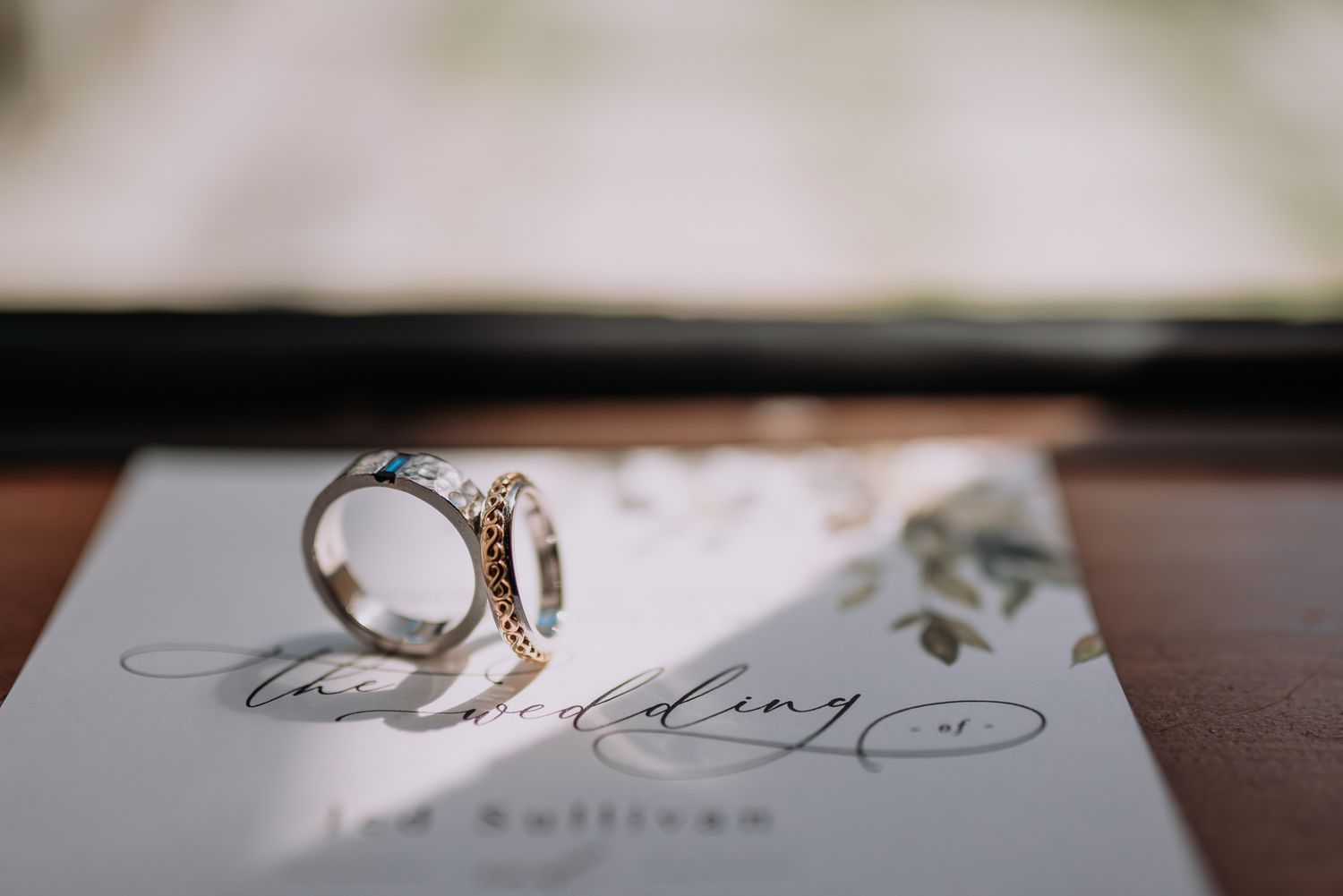 Two elegant wedding rings resting on a calligraphy wedding invitation card near a sunlit window taken by a Melbourne wedding photographer.