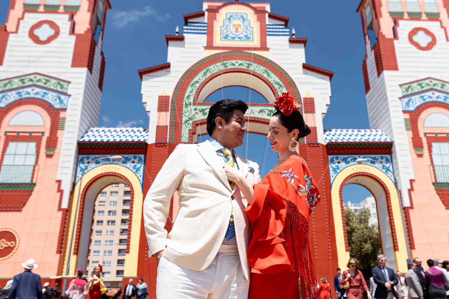 Couple in festive attire at a sunny outdoor fair, man in white suit, woman in red flamenco dress.