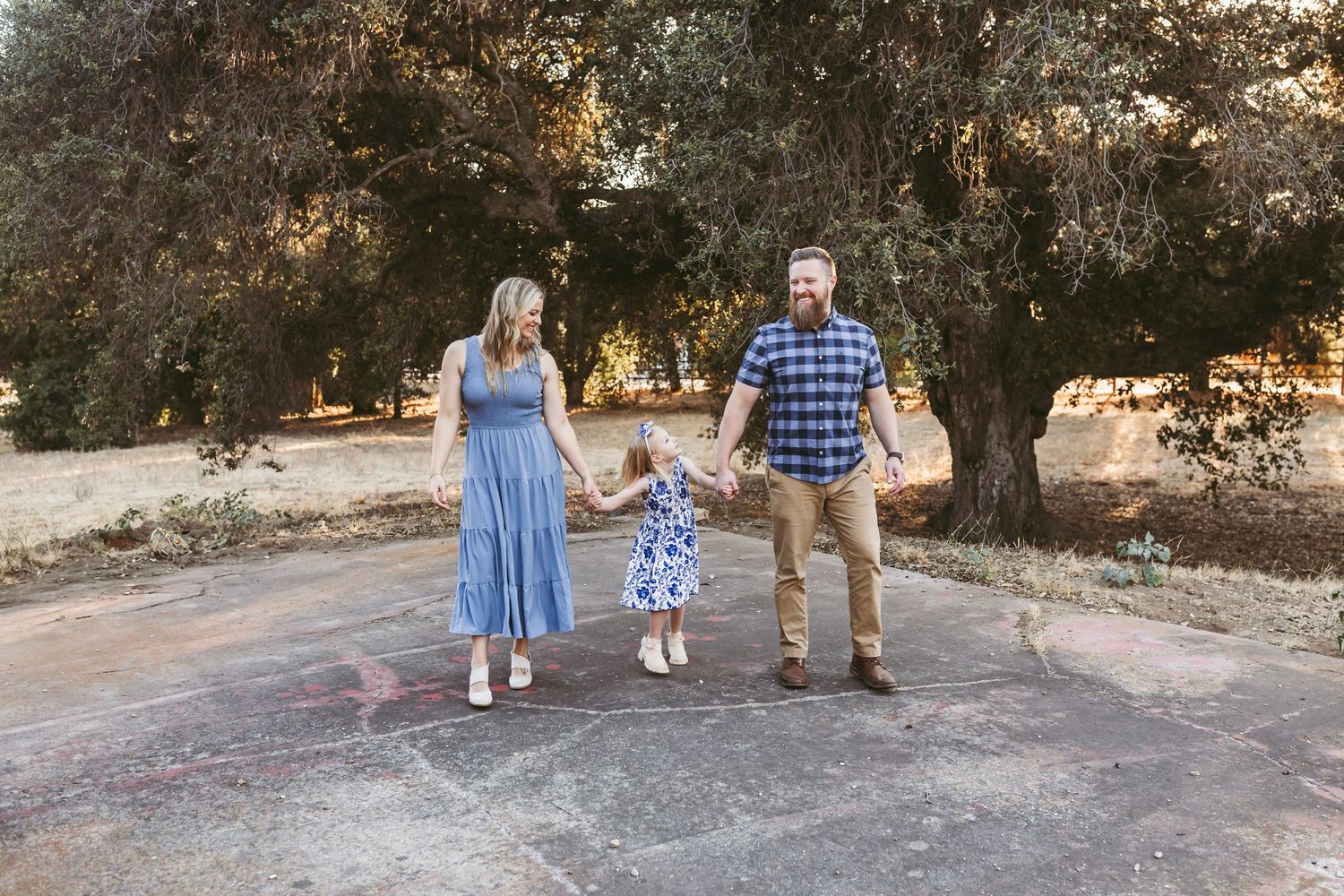 Family of three holding hands and swinging toddler on a sunny tree-lined path at a park in yucaipa, ca