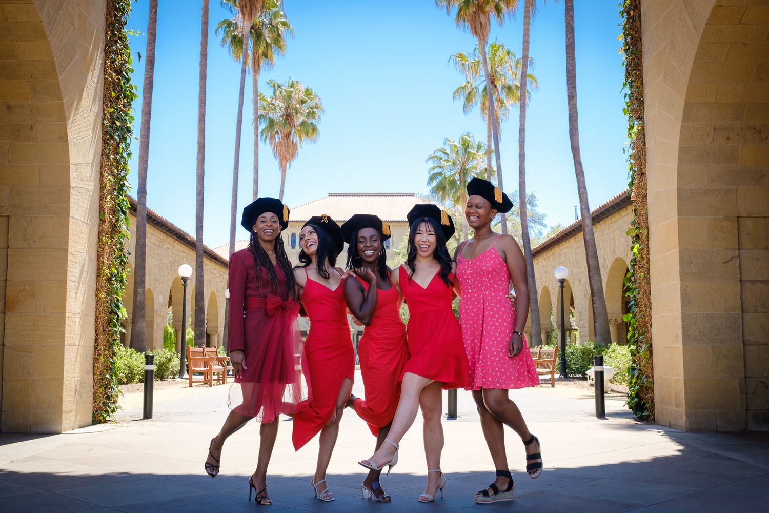 Five women in red dresses and black hats pose confidently outdoors near palm trees and an elegant building.