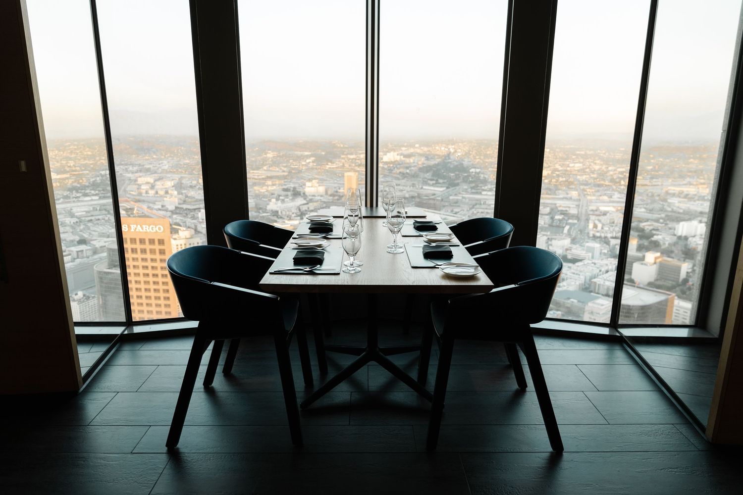 Elegant dining table with black chairs set for four overlooking a stunning city skyline at sunset.