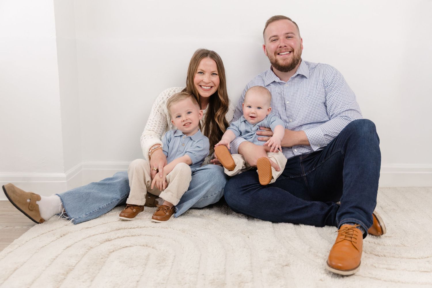 Happy family of four sitting on floor, parents smiling with two young toddler boys in matching denim outfits.