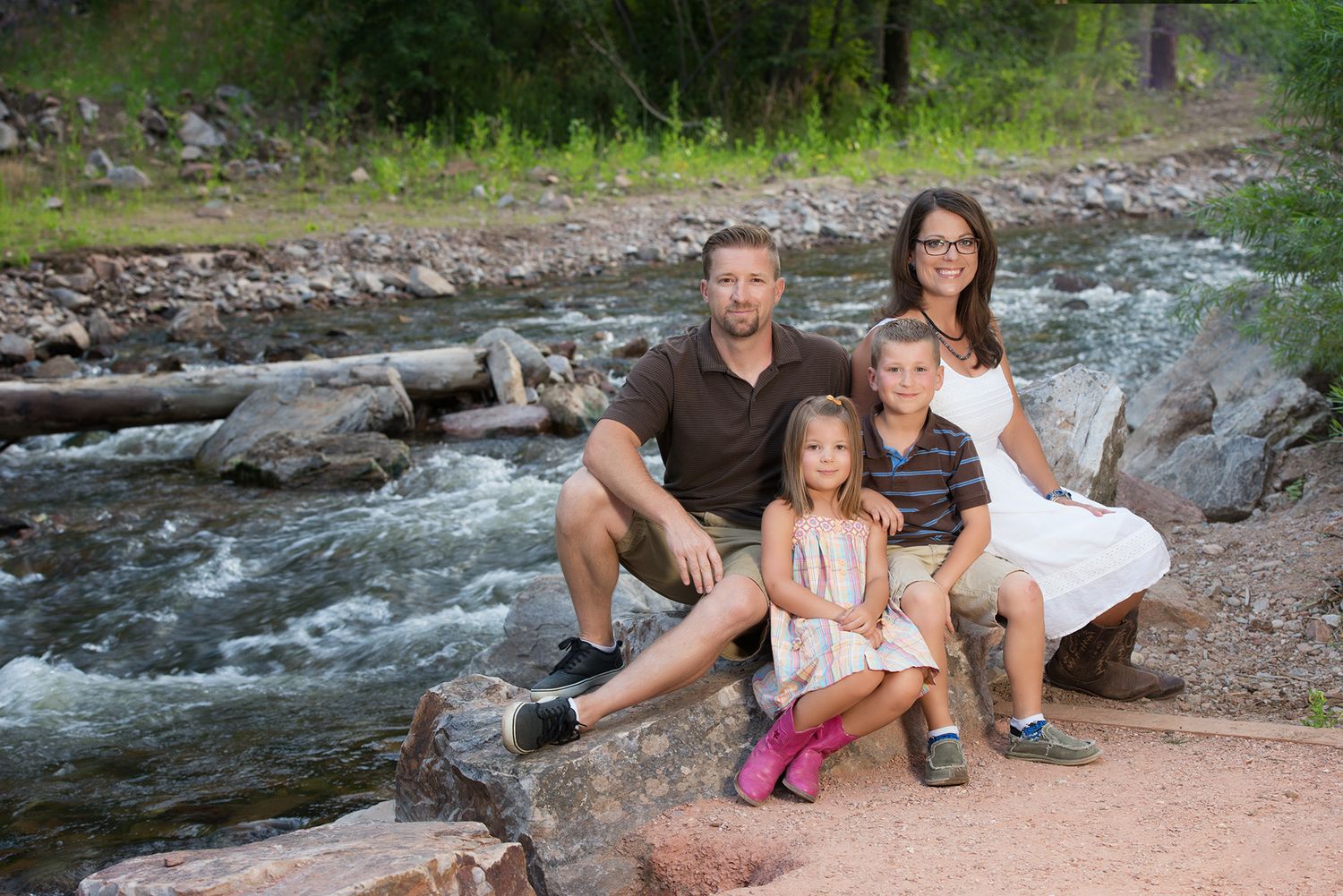 Family of four poses on rocks beside a rushing mountain stream surrounded by lush green trees.