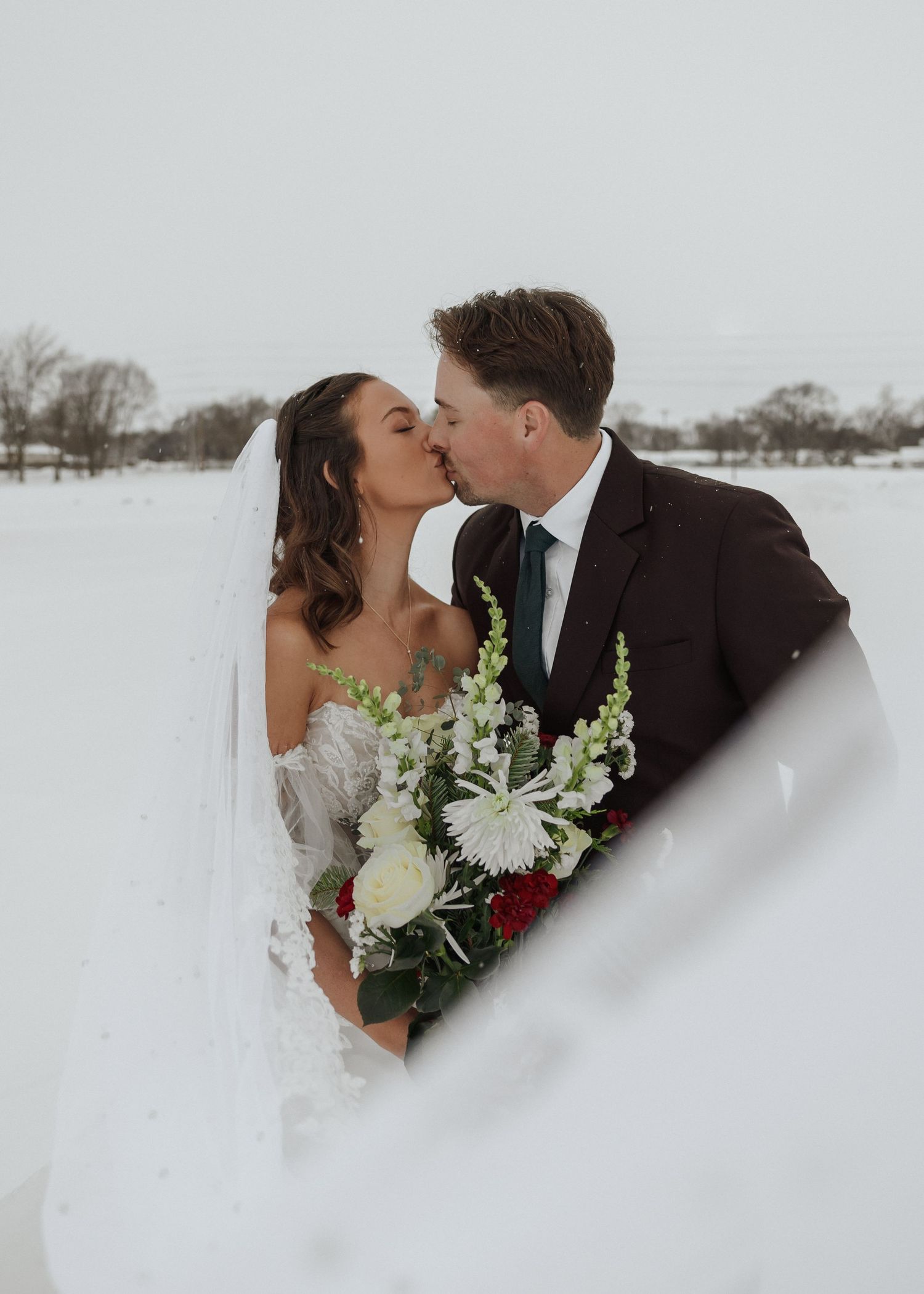 Bride and groom share a kiss in snowy winter landscape, bride holding red and white floral bouquet.