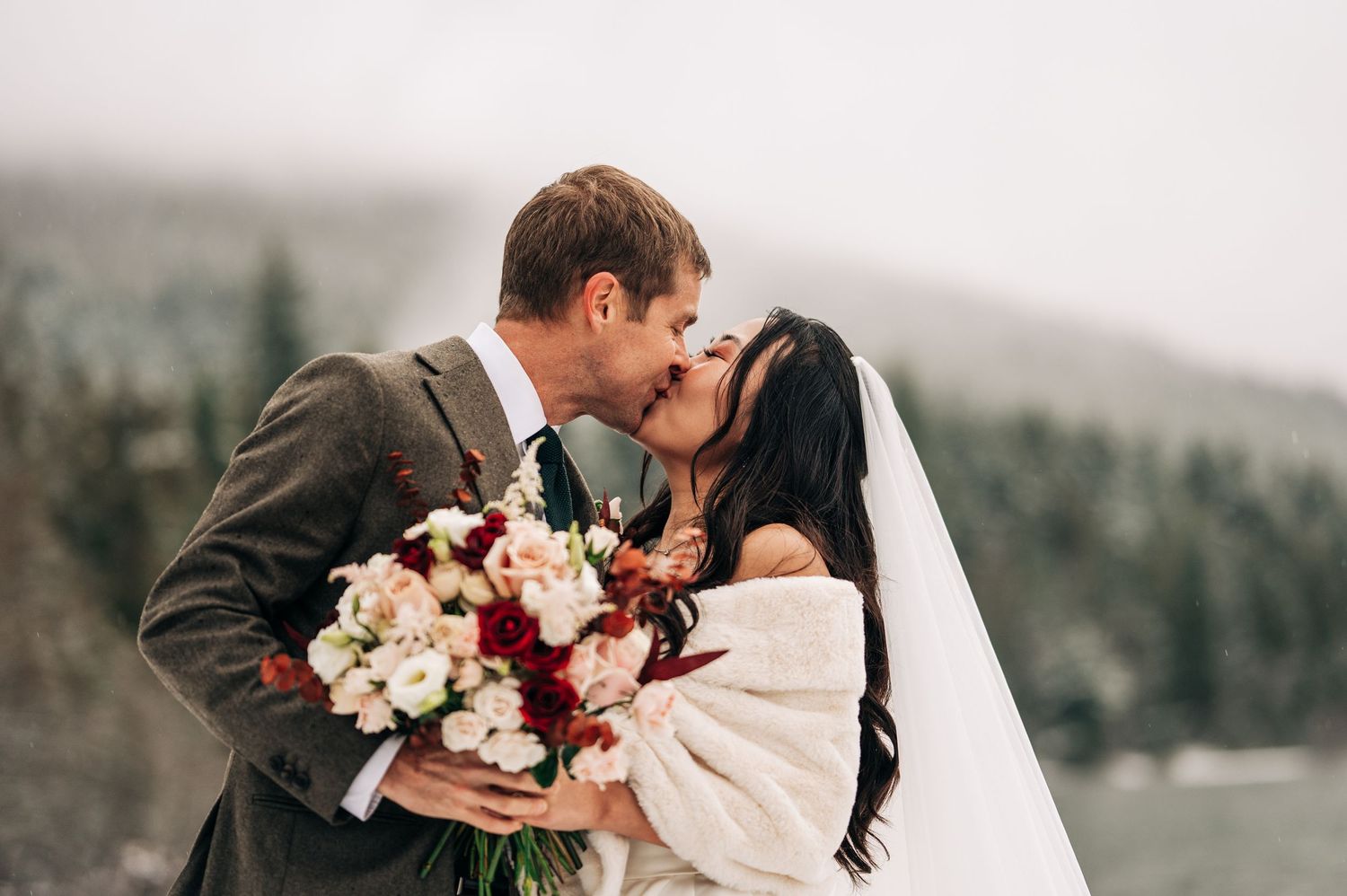 A romantic winter wedding scene with snow-covered mountains and evergreen trees in the background.