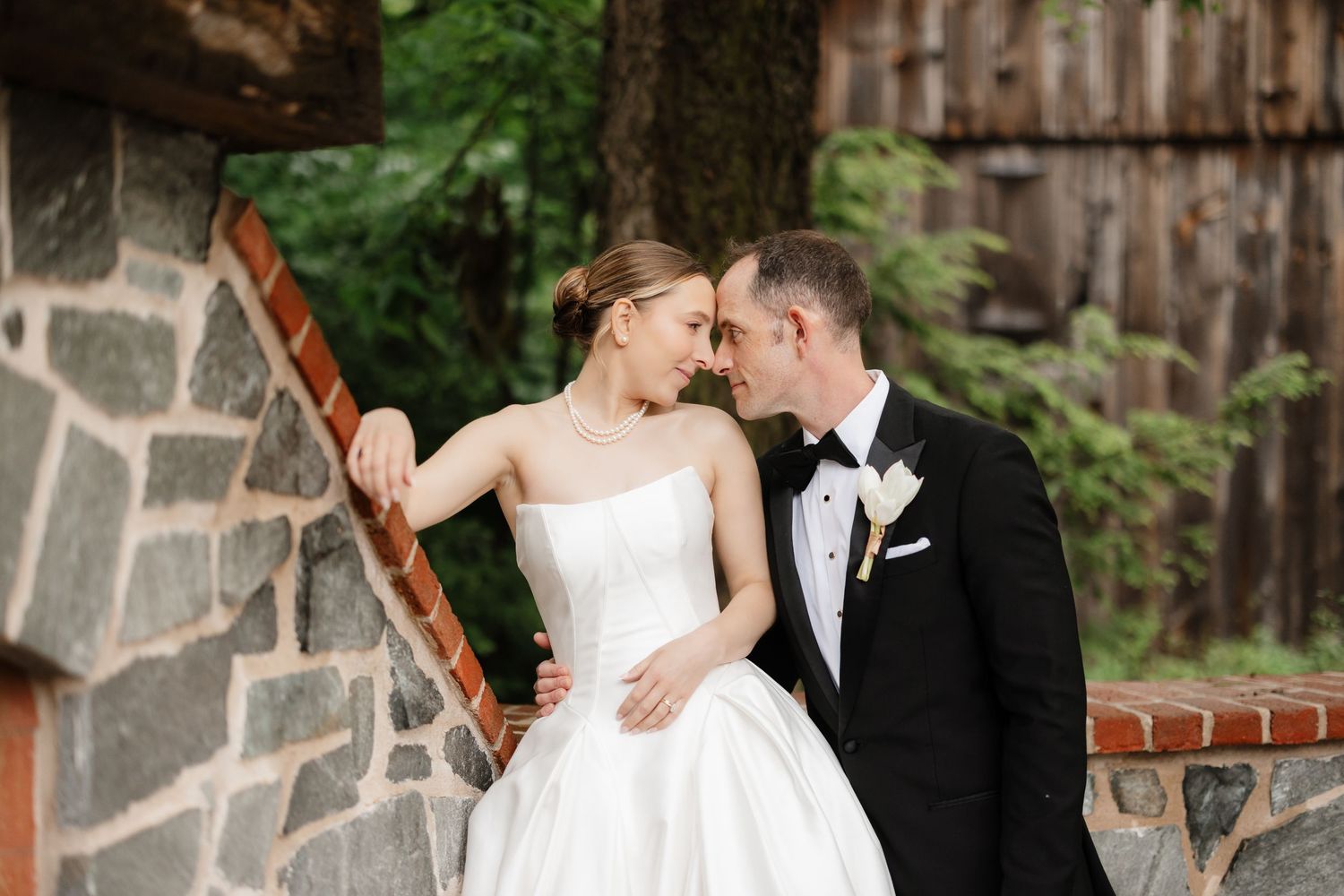 Bride and groom share an intimate moment leaning against a rustic stone wall surrounded by green trees.