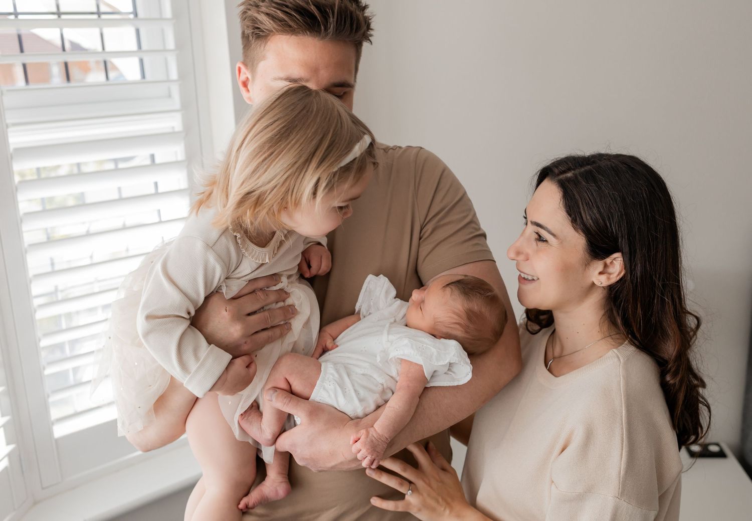 A young family with a newborn baby and toddler sharing a tender moment by a bright window indoors.