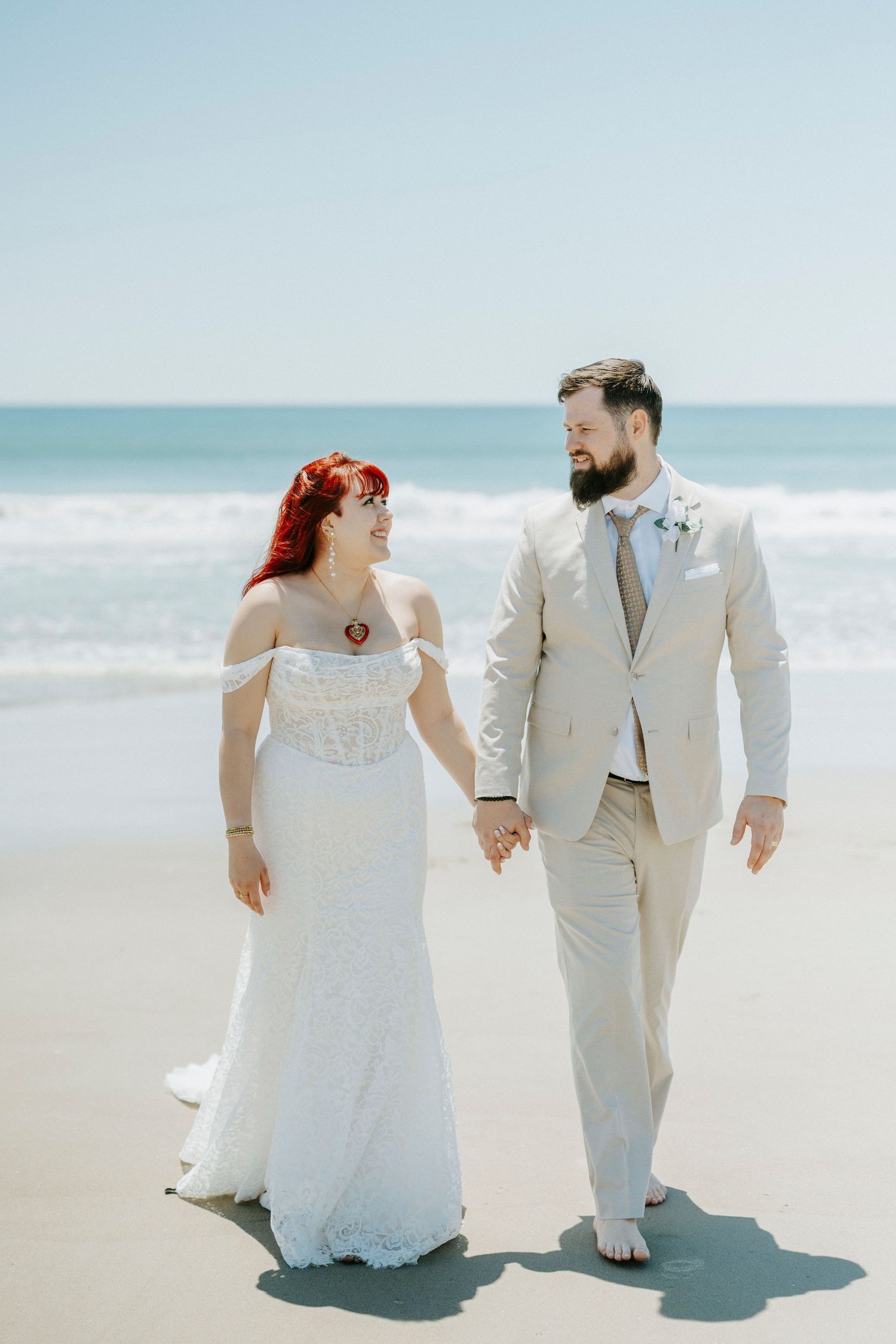 Bride and groom walk hand in hand on a sunny beach, she in white lace gown, he in beige suit.