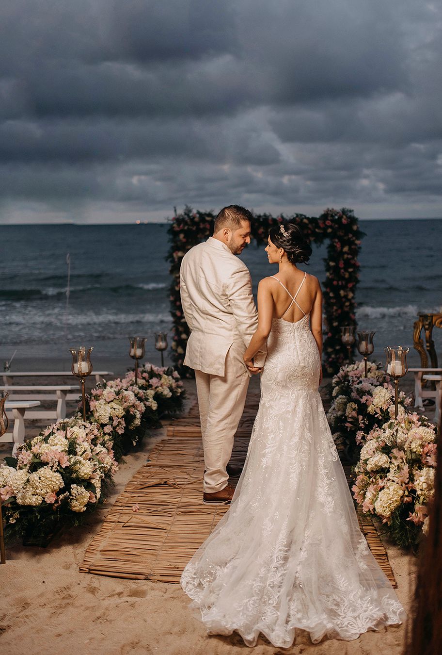 Bride and groom hold hands at romantic beach wedding ceremony with floral arch and stormy ocean backdrop.
