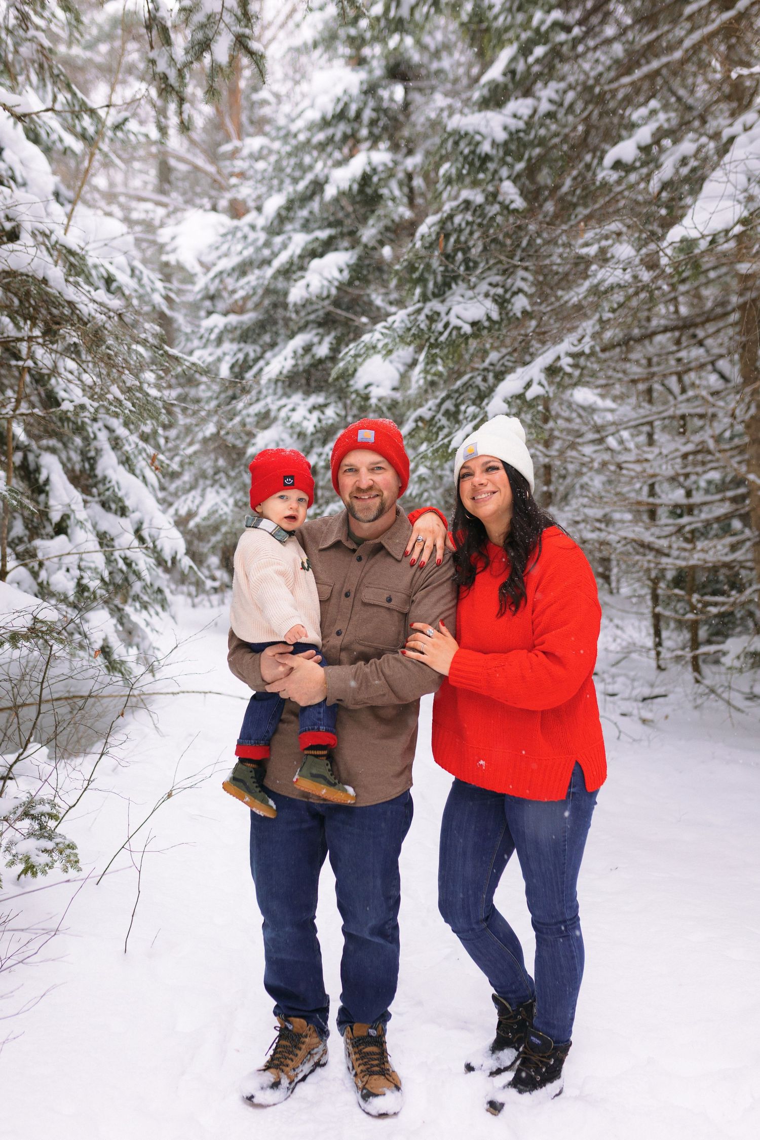 A family wearing winter clothing poses together in a snowy forest setting with snow-covered evergreen trees in the background.