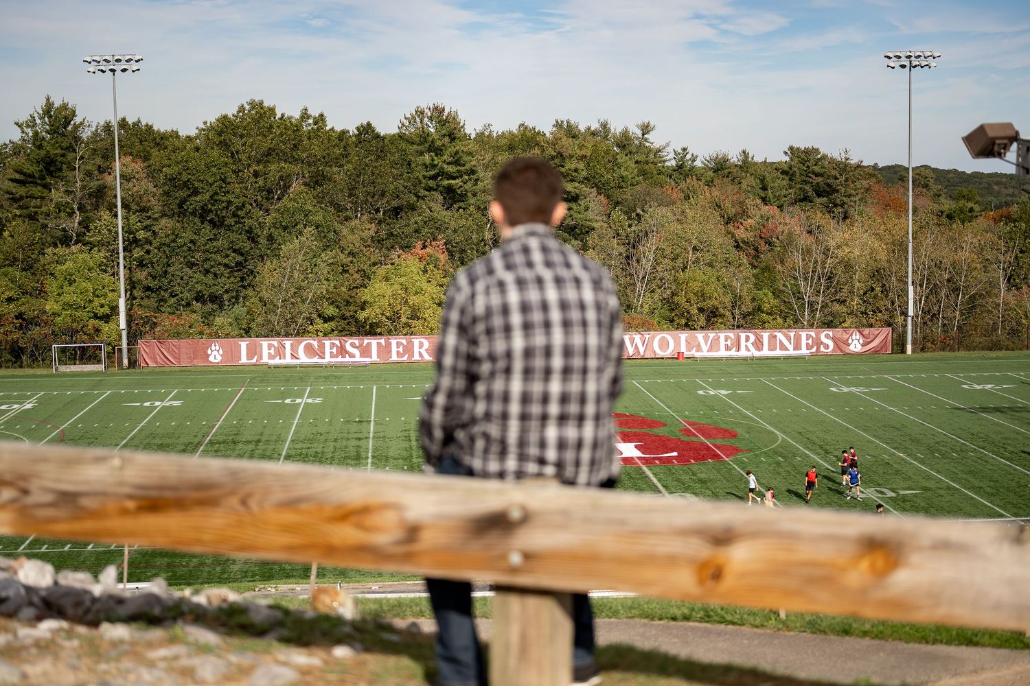 A panoramic view of an empty football field with stadium lights and trees in the background on a sunny day.