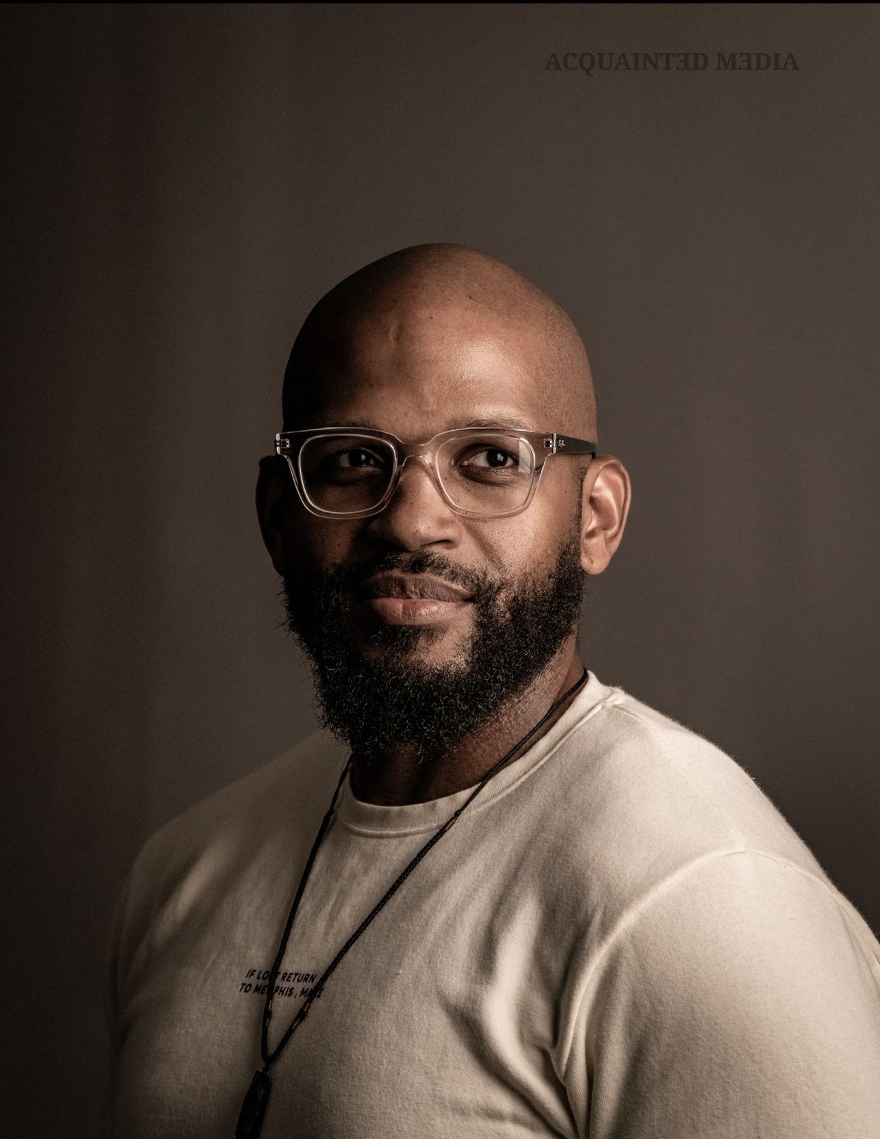 Bald Black man with beard and clear-framed glasses wearing a beige t-shirt and necklace, professional portrait photo.