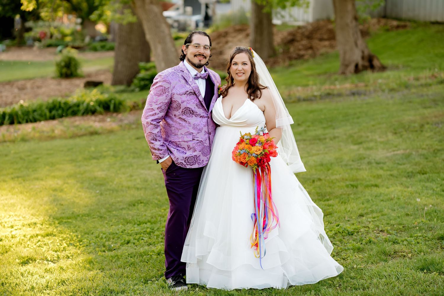 Bride in white gown holding colorful bouquet stands with groom in purple suit on green lawn at outdoor wedding.