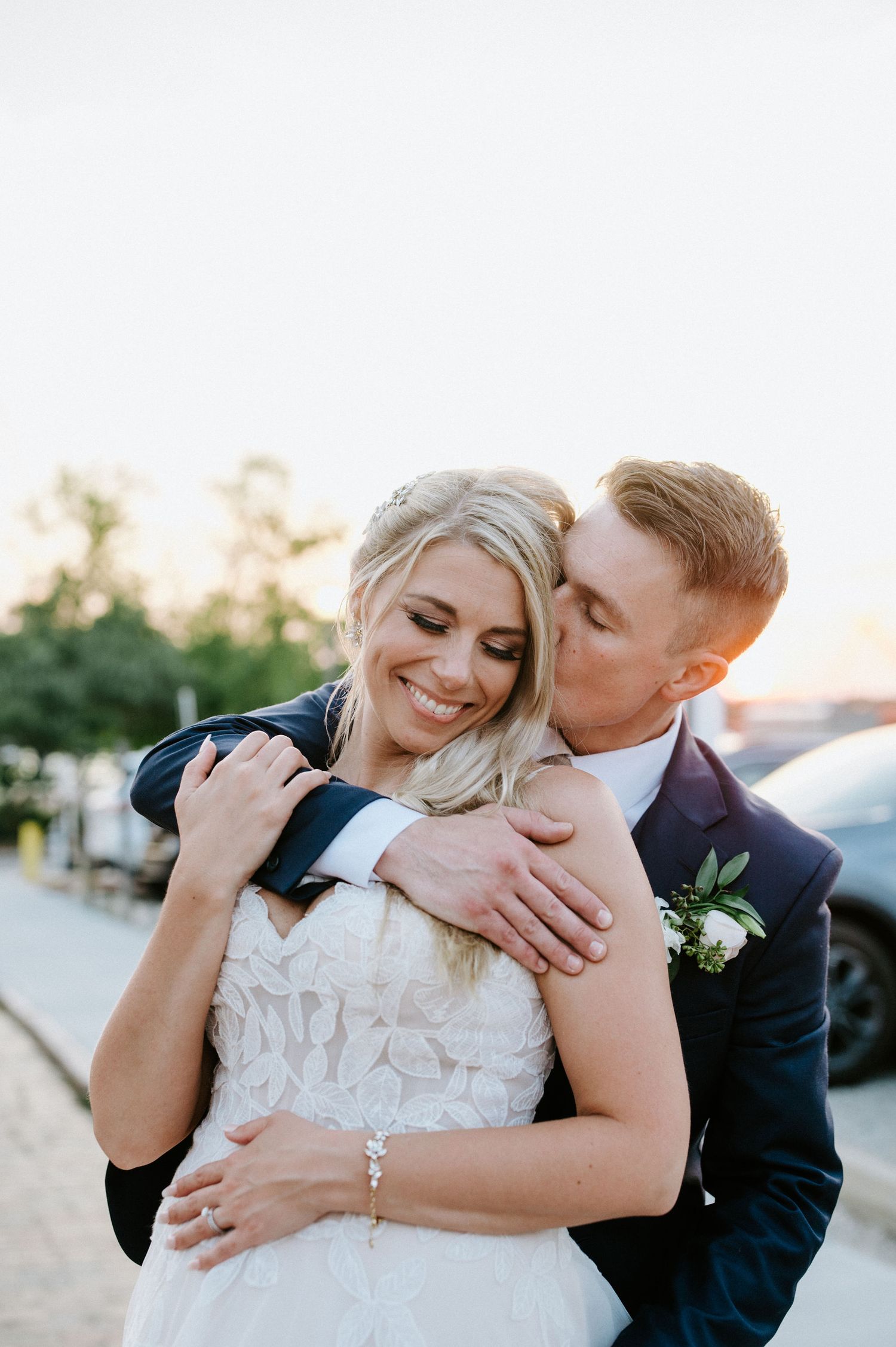 A romantic wedding couple embraces outdoors during golden hour, with the bride in white and groom in navy suit.