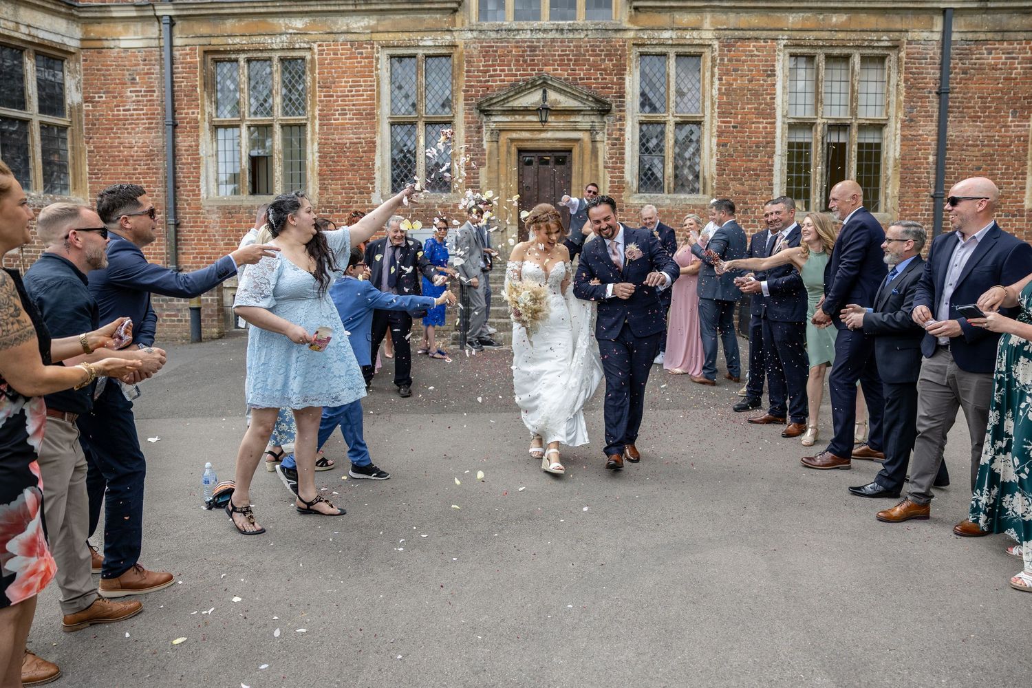 Wedding guests line up outside a historic brick building to shower the newlyweds with confetti and rice.