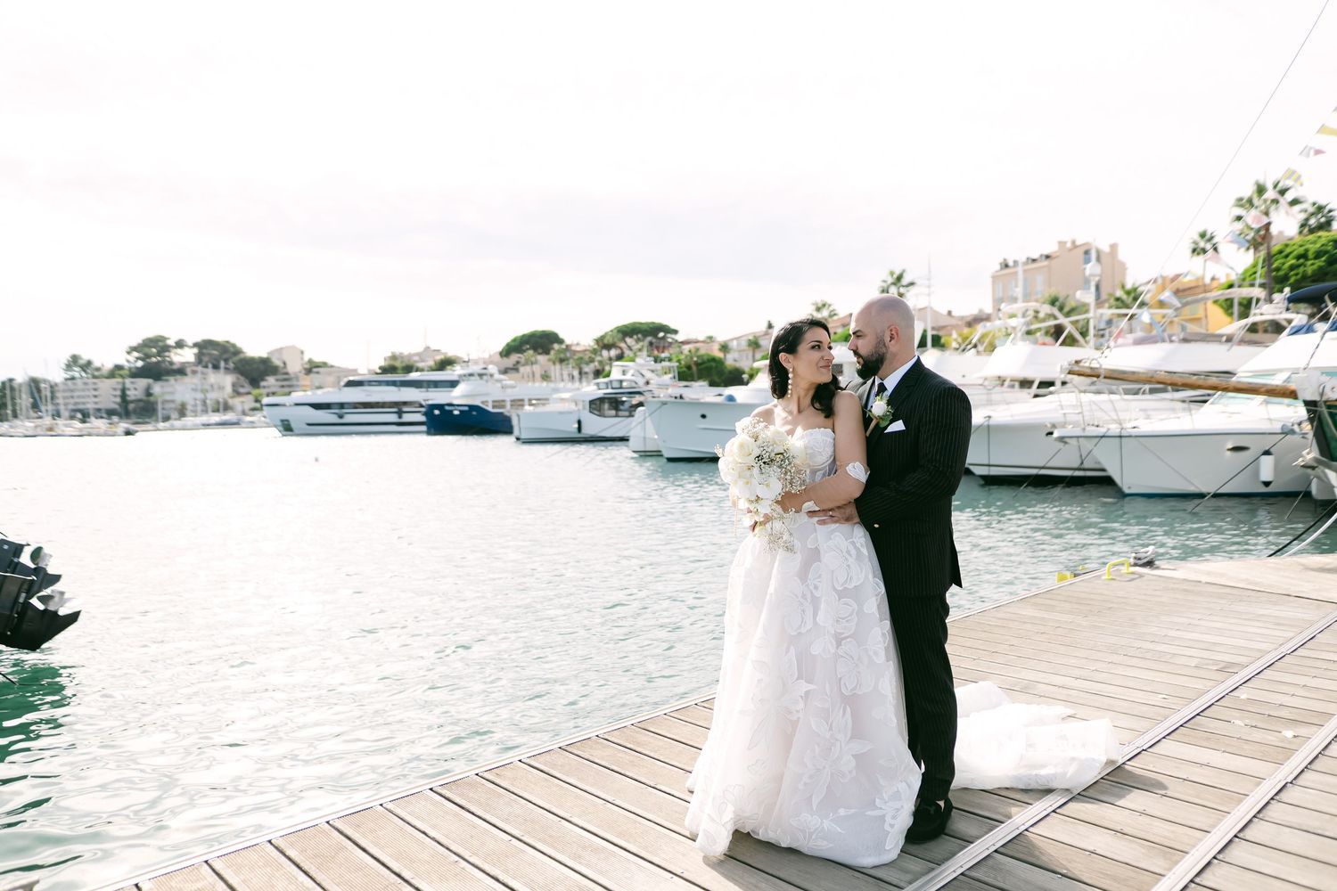 A couple in formal wedding attire embraces on a wooden dock with luxury boats and marina in the background.