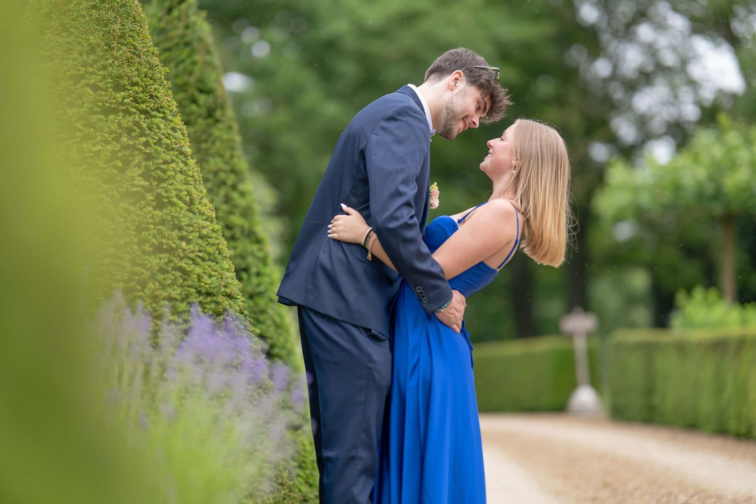 A couple in formal attire embraces in a lush garden setting with vibrant green hedges and trees.