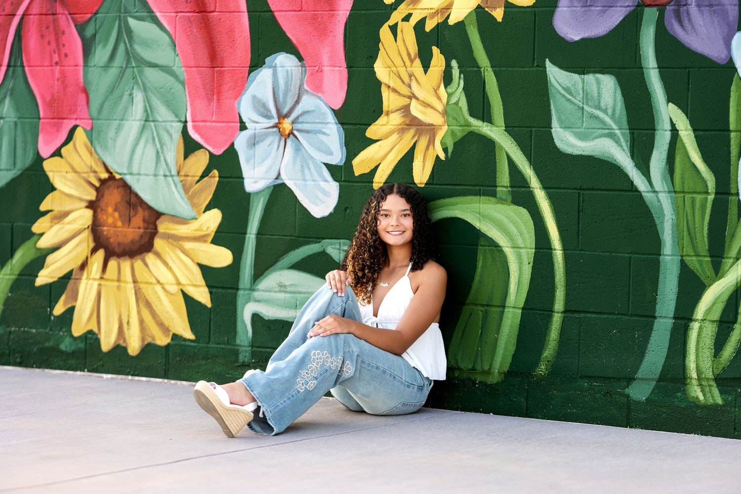 Young woman with curly hair sits smiling against a vibrant floral mural in jeans and white top.