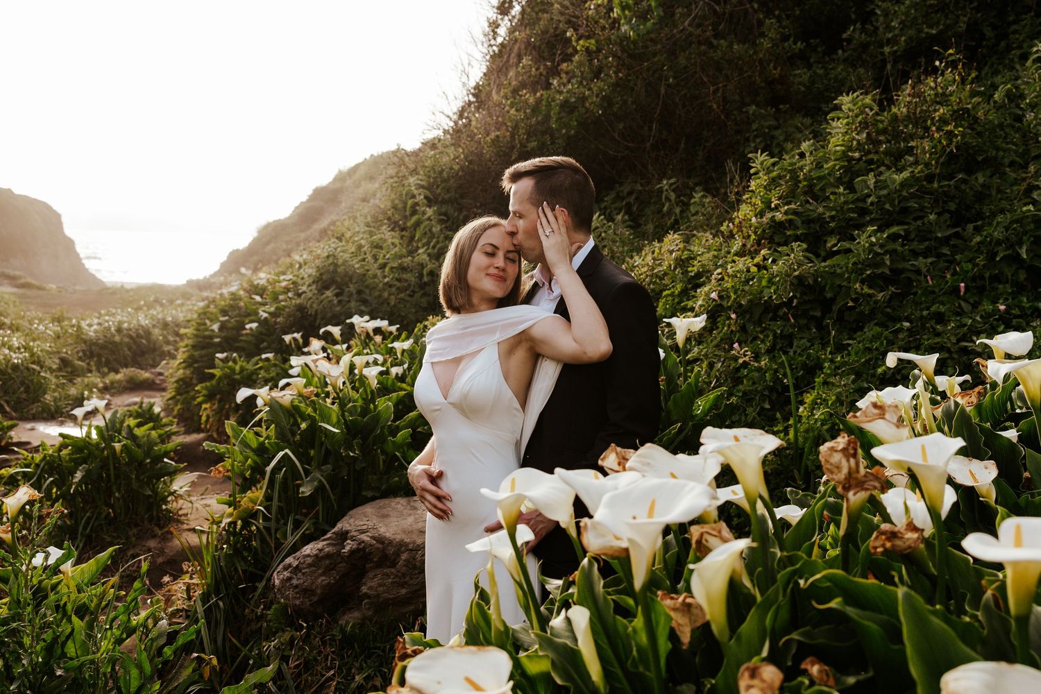 bride and groom snuggling while groom kisses bride's forehead while they're standing in a valley of calla lilies in Carmel-by-the-Sea