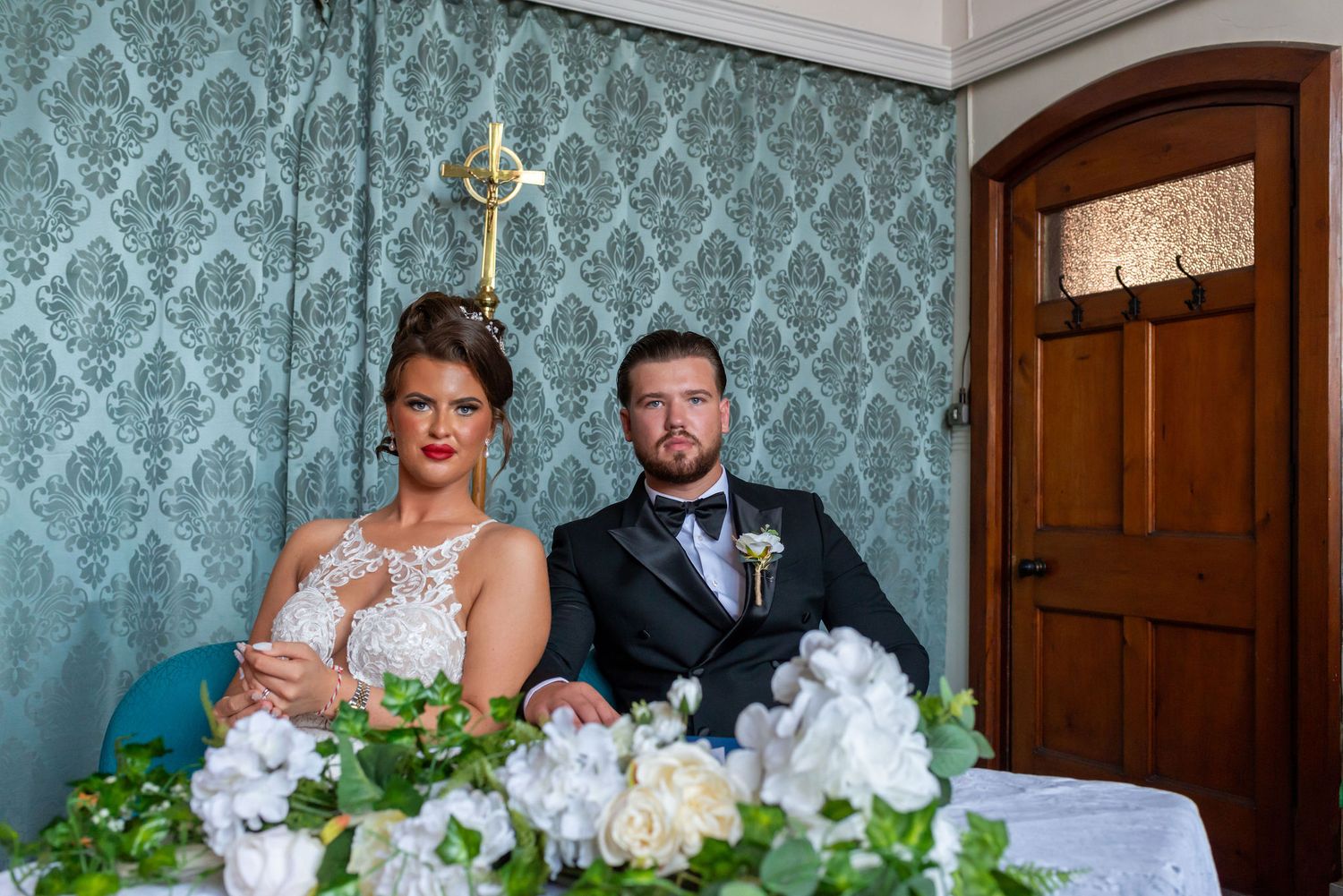 Bride and groom seated at wedding reception table with white floral centerpiece and elegant teal damask wallpaper.
