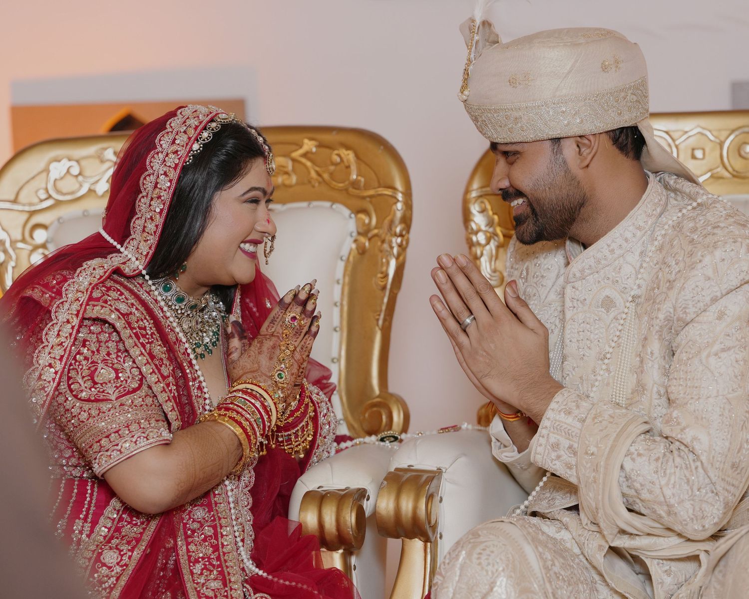 Indian bride in red lehenga and groom in cream sherwani exchange Namaste greetings during traditional Hindu wedding ceremony.