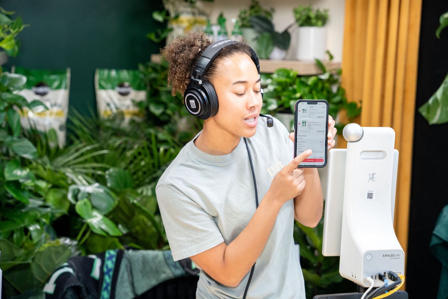 Young woman with headphones holding smartphone surrounded by lush green plants in a modern indoor setting.