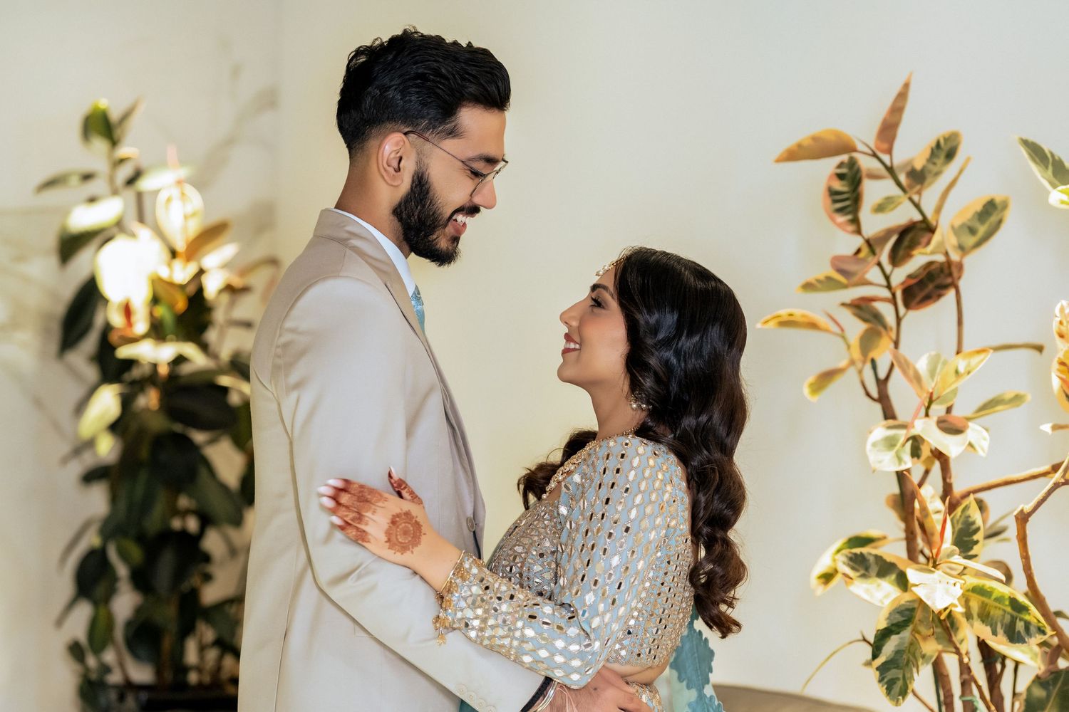 South Asian couple in elegant attire sharing a romantic moment, bride with mehndi henna, surrounded by lush greenery.