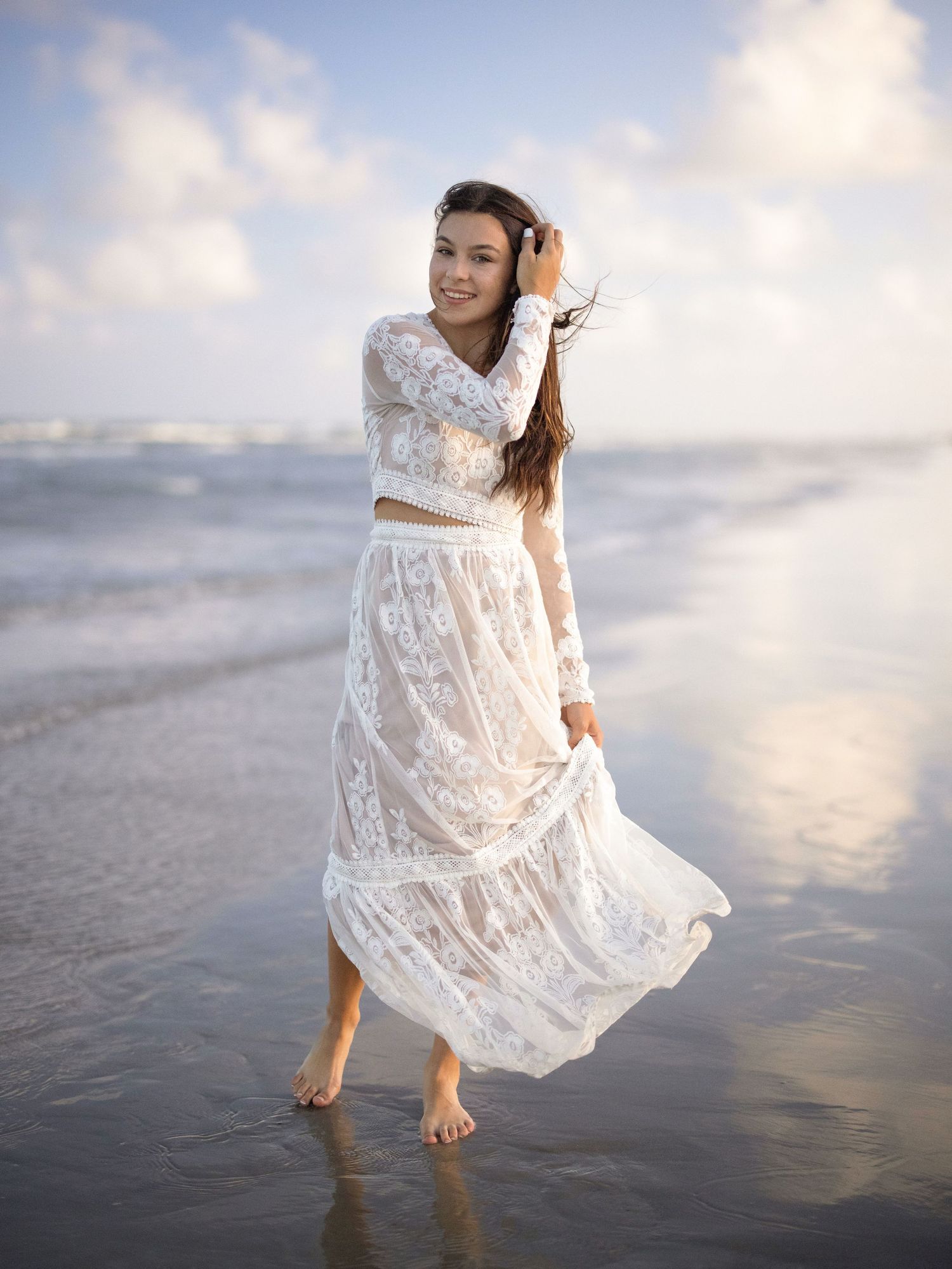 Young woman in white lace two-piece dress standing barefoot on a beach with ocean waves and cloudy sky.