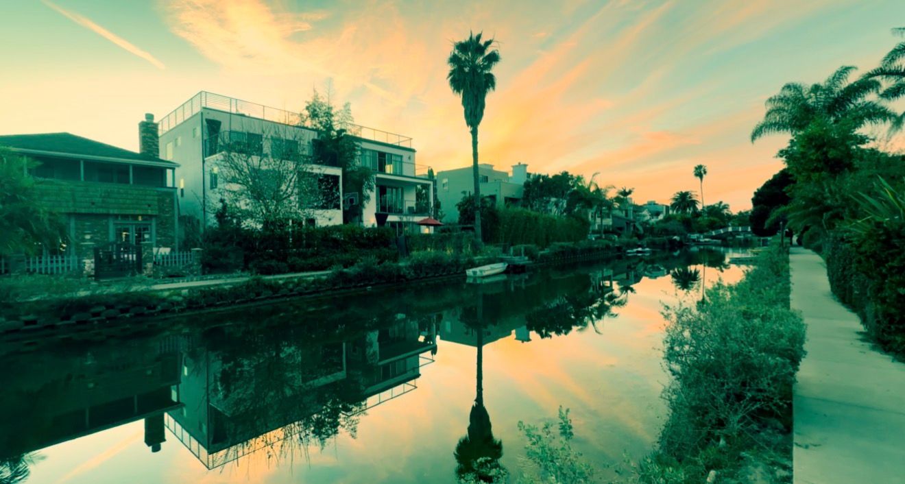 A panoramic view of a waterfront neighborhood with palm trees and houses reflected in calm waters at sunset.
