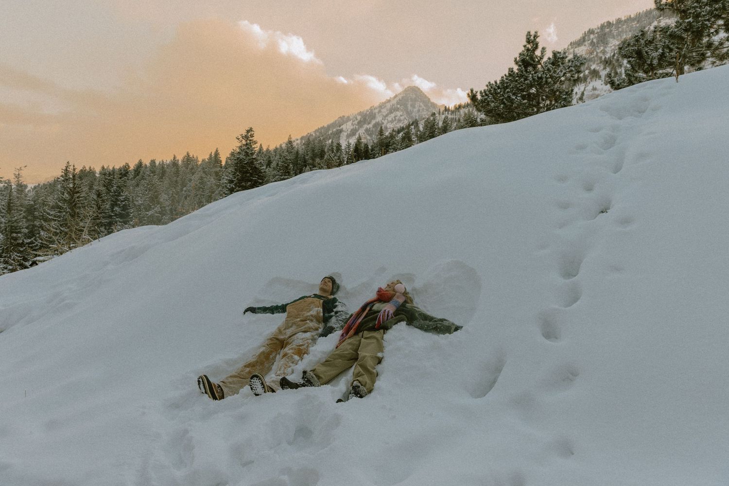 Couple playfully rolling down a snowy mountain slope during a winter photoshoot.